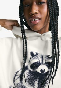 White hoodie featuring a detailed raccoon illustration in black, with braided hair partially visible, emphasizing the clothing's design.