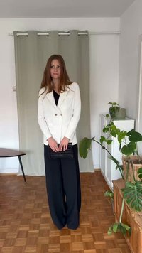 Blazer branco com bainhas, com dois botões, combinado com uma blusa preta e calças largas pretas. A segurar uma clutch preta texturizada. Chão de madeira.