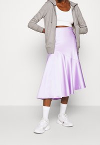 Jupe midi en satin violet avec un design asymétrique, associée à un sweat à capuche gris zippé et des baskets blanches, sur un fond neutre.