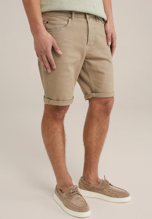 Homme portant un short beige retroussé et des chaussures bateau en daim assorties à lacets, debout devant un fond neutre.
