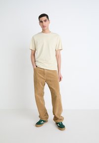 Beige katoenen t-shirt, ontspannen fit, gecombineerd met lichtbruine corduroy broek; model draagt groene sneakers met witte accenten. Minimalistische stijl.