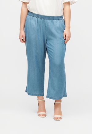 Kvinde iført lyseblå vidde-benet cropped jeans med elastisk talje og hvide åbne tå ankelstropper med hæle.