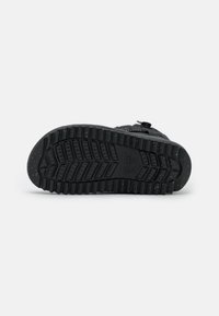 Semelle de chaussure noire avec un profond relief, présentant des motifs en relief, une texture mouchetée et un logo circulaire sur le côté. Bords lisses et forme arrondie.