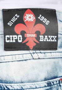 Black leather label featuring a red fleur-de-lis design, white text "CIPO DNM BAXX," and "SINCE 1996" against light blue denim fabric.