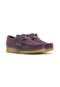 Purple suede shoes with a round toe, laced with multicolored cords, and a light brown rubber sole. Features a signature label on the side.