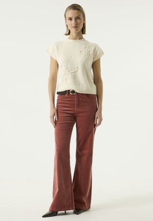 Frau steht und trägt einen cremefarbenen Kurzarmpullover mit Blumendetails, rostfarbene weit geschnittene Hose, schwarzen Gürtel und schwarze spitze Schuhe.