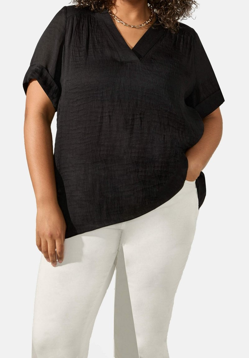Blusa nera con scollatura a V, maniche corte arrotolate e tessuto testurizzato. Abbinata a pantaloni bianchi, in un taglio rilassato.