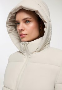 Beżowa kurtka puchowa z dużym, regulowanym kapturem. Posiada pikowaną strukturę i zamek błyskawiczny, z gładkim, błyszczącym wykończeniem materiału.