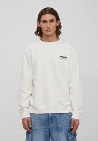 Biała bluza z okrągłym dekoltem, z ribbowanymi mankietami i brzegiem, z małym czarnym logo "VENICE" na klatce piersiowej, wykonana z miękkiego materiału. Casualowy fason.