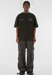Tricou din bumbac negru cu mâneci scurte, având un logo grafic pe piept, asociat cu pantaloni cargo gri închis cu multiple buzunare.