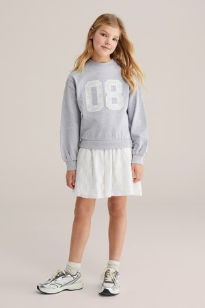 Fiatal lány hosszú szőke hajjal, szürke pulóvert visel „08” felirattal, fehér csipkerokát, világos zoknit és ezüst sportcipőt, semleges háttéren áll.