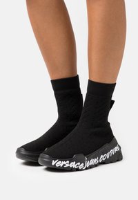 Zapatos de estilo calcetín negros con un upper texturizado, puño elástico y una suela gruesa con una marca impresa en blanco a lo largo del lado.