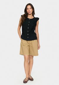 Top senza maniche nero con parte anteriore a balze e chiusura con bottone, abbinato a pantaloni corti beige e scarpe con motivo. Design semplice e pulito.
