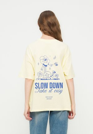 Person med langt brunt hår som har på seg en kremfarget T-skjorte med blå snegle, blomst, bie-grafikk og teksten "SLOW DOWN Take it easy" på ryggen.
