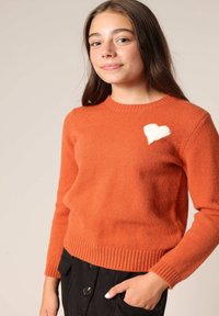Pull en laine orange avec un col rond, des poignets et un ourlet côtelés, comportant un patch en forme de cœur blanc sur la poitrine gauche. Texture douce.