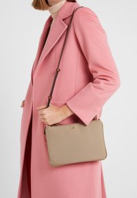 Borsa a tracolla beige con una texture liscia, forma rettangolare slim, accessori in oro e tracolla regolabile marrone, appoggiata su un cappotto rosa.