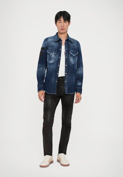 Junger Mann steht da und trägt eine verwaschene blaue Jeansjacke über einem weißen Hemd, schwarze schmale Hosen und weiße Sneakers vor einem einfarbigen Hintergrund.