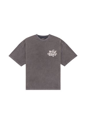 Femme portant un t-shirt gris surdimensionné avec un imprimé blanc, un pantalon noir à jambes larges, et une chemise à carreaux bleue drapée sur une épaule.