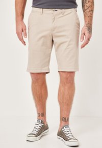 Beige Baumwollshorts mit geradem Schnitt, seitlichen Taschen und einem Knopf am Bund. Kombiniert mit grauen Sneakers mit weißen Sohlen und Schnürsenkeln.
