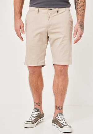 Beige Baumwollshorts mit geradem Schnitt, seitlichen Taschen und einem Knopf am Bund. Kombiniert mit grauen Sneakers mit weißen Sohlen und Schnürsenkeln.