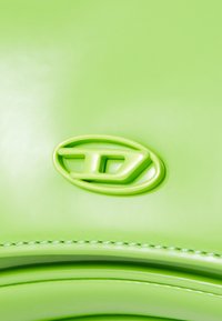 Zelena usnjena torba z izrazitim ovalnim logom. Površina je gladka z sijočim zaključkom in kontrastnim belim šivom ob robu.