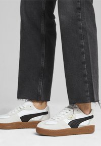 Baskets en daim blanc et noir avec semelles en caoutchouc marron ; caractéristiques : design à lacets et accents de logo sur le côté. Portées avec un jean noir effiloché.