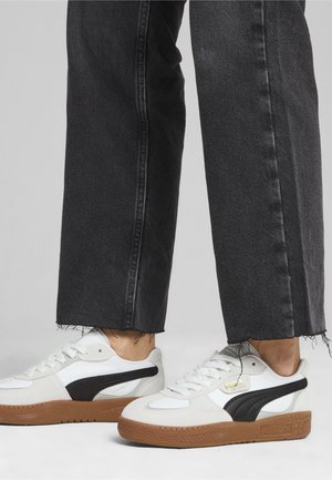 Białe i czarne zamszowe sneakersy z brązowymi gumowymi podeszwami; cechy obejmują sznurowanie i logo na boku. Nosi się je z przetartymi czarnymi dżinsami.