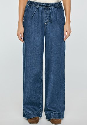 Kobieta ubrana w luźne, wysokie spodnie z niebieskiego denimu z troczkiem i brązowe buty, stojąca na jednolitym tle.