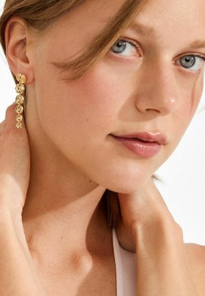 Kullast tooniga tilgavad kõrvarõngad, millel on spiraalkuju ja mitu omavahel linked ringi. Mudelil on hele nahk ja sirged, õlgadeni ulatuvad juuksed.