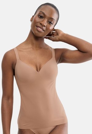 Woman wearing a nude-colored camisole and matching underwear, smiling with one hand resting on the back of her neck against a plain background.