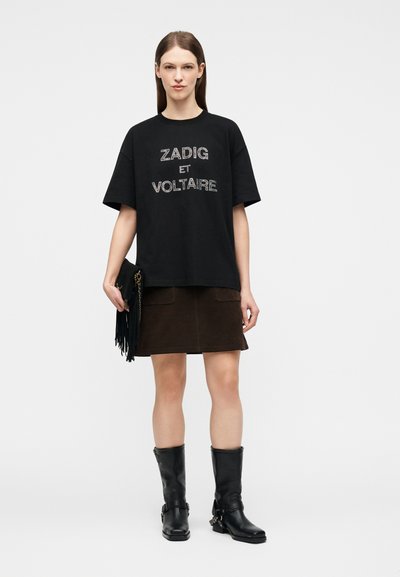 Νεαρή γυναίκα στέκεται φορώντας μαύρο φαρδύ T-shirt με το κείμενο "Zadig et Voltaire", σκούρα μίνι φούστα, μαύρες μπότες μέχρι τη μέση της γάμπας και κρατώντας τσάντα φάκελο με κρόσσια.