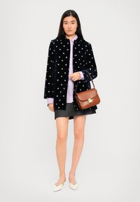 Frau in einer schwarzen Blumenjacke, lila Hemd, grauem Rock und weißen spitzen Schuhen, die eine braune Lederschultertasche trägt.