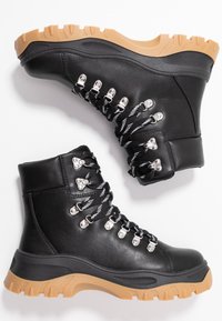 Botas de senderismo de cuero negro con un acabado texturizado, ojales de metal y cordones con un patrón en blanco y negro. Suela gruesa de goma de color beige.