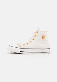 Białe trampki typu high-top na płótnie z złotymi oczkami, czarną gumową podeszwą i haftowanym sercem po zewnętrznej stronie.