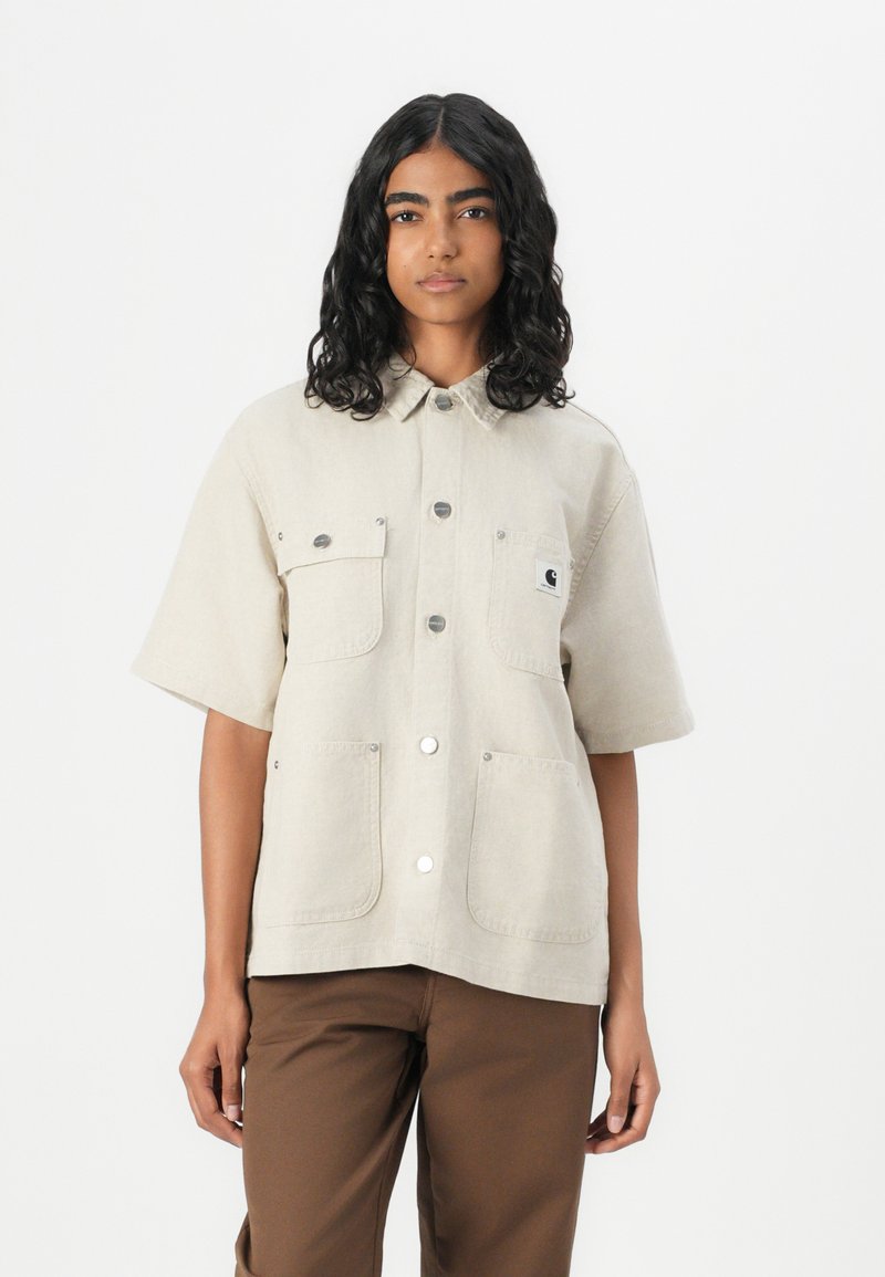 Beige short-sleeve shirt with a button-down collar, two chest pockets, silver buttons, and a brand logo patch on the left side.