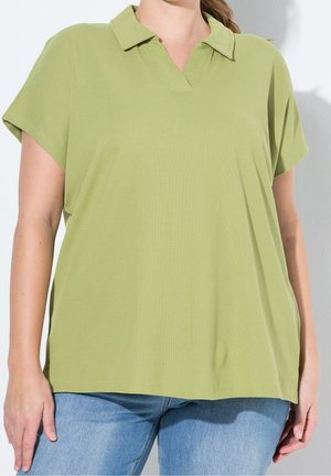 Osoba nosząca luźną jasnooliwkową koszulkę polo z krótkim rękawem i niebieskie dżinsy, stojąca na jednolitym jasnym tle.