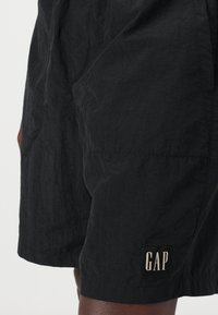 Sorte shorts i et tekstureret stof, med en sidelomme og et rektangulært GAP-logo i hvidt på sort baggrund.