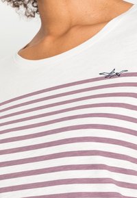 T-shirt a righe con base bianca e strisce viola alternate, con colletto rotondo e un piccolo dettaglio ricamato di un uccello.