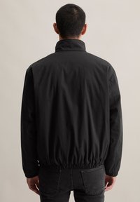 Man in een zwarte jas en zwarte jeans, met zijn rug naar de camera tegen een effen lichte achtergrond.