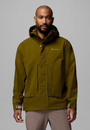 Mand iført en olivengrøn Columbia hættejakke med store forlommer og beige bukser, stående mod en grå baggrund.