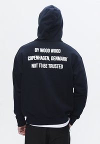 Tengerészkék kapucnis pulóver, elülső kenguruzsebbel. A hátán nagy, fehér felirat áll: "BY WOOD WOOD COPENHAGEN, DÁNIA, NEM MEGBÍZHATÓ".