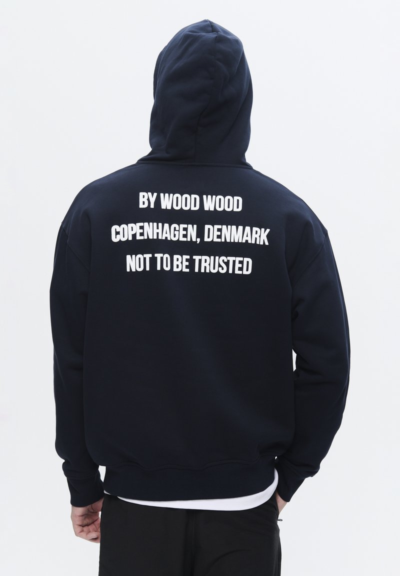 Tengerészkék kapucnis pulóver, elülső kenguruzsebbel. A hátán nagy, fehér felirat áll: "BY WOOD WOOD COPENHAGEN, DÁNIA, NEM MEGBÍZHATÓ".