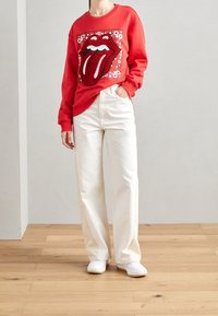 Rotes Sweatshirt mit großem Zungenlogo, kombiniert mit weißen, weiten Hosen und weißen Turnschuhen vor neutralem Hintergrund.