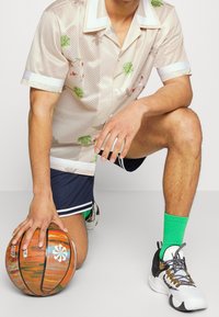 Camisa de malla beige de manga corta con estampados florales en verde y rojo; pantalones cortos azul marino; calcetas deportivas verdes; sosteniendo un balón de baloncesto naranja.