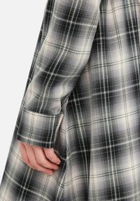 Hånd hvilende ved siden af et sort-hvidt ternet langærmet tøjstykke med synlig manchet og fine syningsdetaljer.