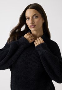Pull noir texturé avec un col haut et des manches larges. Le tissu semble doux et moelleux, offrant un aspect confortable. Pas de motifs ou d'accents visibles.