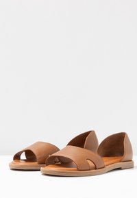 Sandalias de cuero marrón con un diseño minimalista, que presentan dedos al aire y acentos recortados. Suela plana con una plantilla texturizada.
