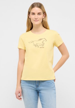 Frau trägt ein gelbes T-Shirt mit schwarzer Kontur eines galoppierenden Mustang-Pferdes und hellblaue Jeans, steht mit einer Hand hinter dem Rücken.