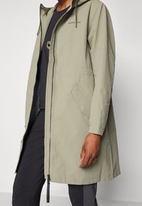 Světlé zelené voděodolné sako s kapucí, zipovým uzávěrem a dvěma předními kapsami; má volný, k délce kolen navržený střih a elastické manžety.