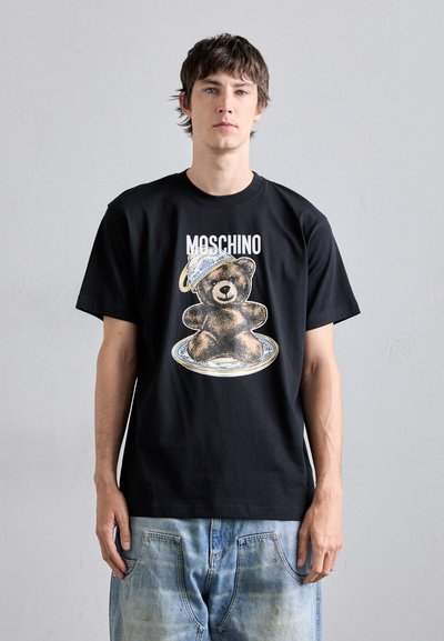 Musta puuvillapaita, jossa on keskellä karhugrafiikkaa, ympäröity koristelevyllä; mukana "MOSCHINO"-teksti ylhäällä.